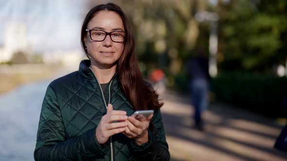 Calm Joyful Woman is Using Smartphone on City Street Smiling and Looking at Camera alt