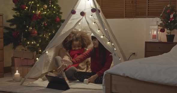Two little girls having video call on tablet computer in decorated teepee tent at home