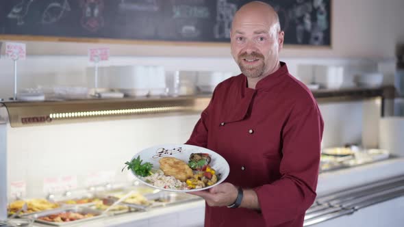 Portrait of Satisfied Bearded Male Cook Showing Plate with Delicious Lunch Looking at Camera Smiling alt