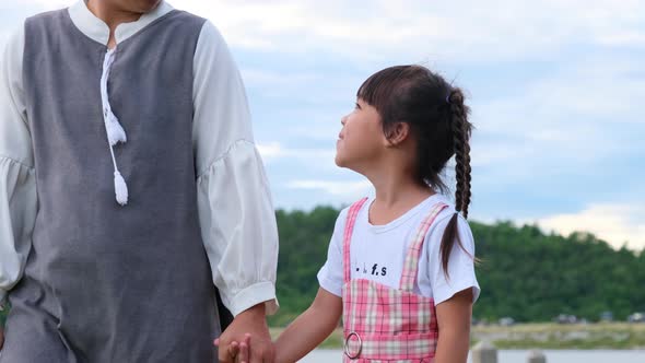 Happy mother walks hand in hand with a little girl and smiles at each other by the lake. alt