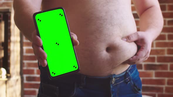 Man Holds Vertically Phone with Chromakey at Level of a Fat Belly with Other Hand Squeezes Full alt