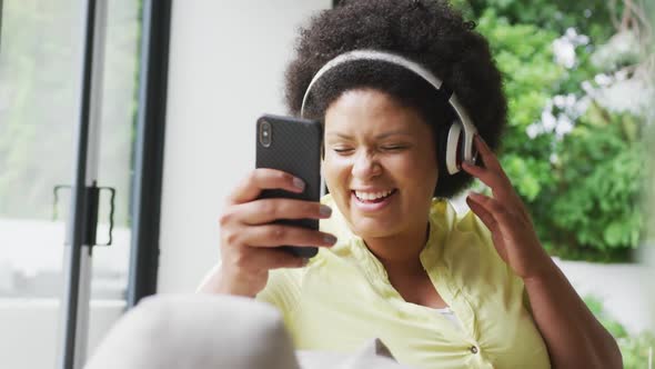 Video of happy plus size african american woman with headphones on sofa, listening to music alt