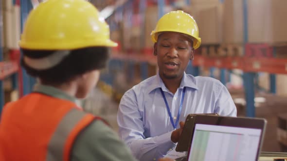 African american male and female workers wearing helmet and using laptop in warehouse alt