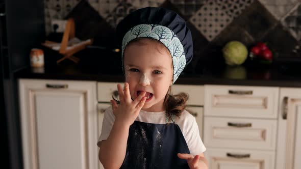 Cute Little Girl Smeared with Flour Licks Her Fingers with Dough in the Kitchen. alt