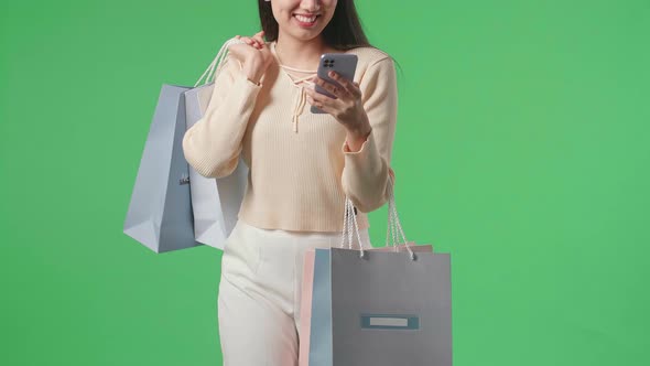 Asian Woman Holding Shopping Bags Use Mobile Phone And Smile While Walking In Front Of Green Screen alt