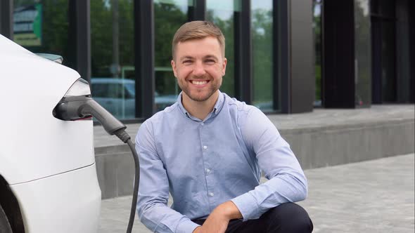 Hansome Guy Sitting Near His New Modern Electric Car While Car is Charging at the Charging Station alt