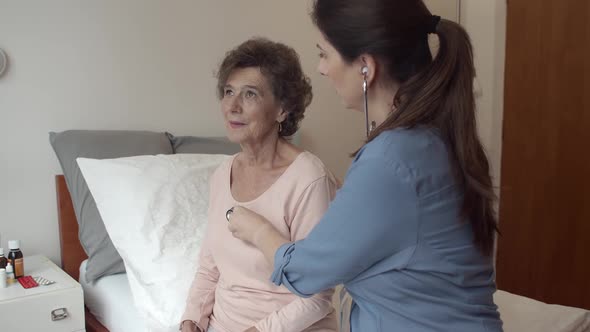 Home Caregiver Listening For Breathing of Elderly Woman Sitting on Bed