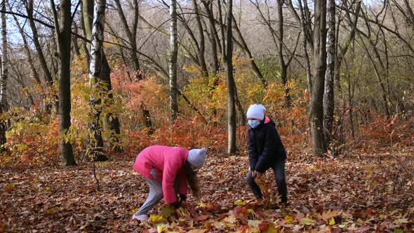 Two girls in medical masks throw fallen yellow leaves in the Park on an autumn day