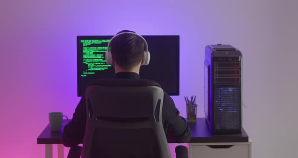 Rear View of Man Programmer in Headphones Coding Network Security Software Indoors in Neon Lights at alt