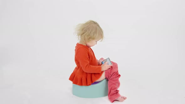 Disheveled Child Sits on Children's Potty Using Smartphone on White Background