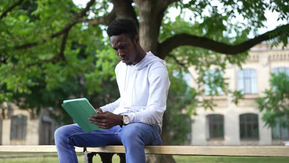 Confident Focused African American Man Surfing Internet on Tablet Drinking Morning Coffee Sitting on alt
