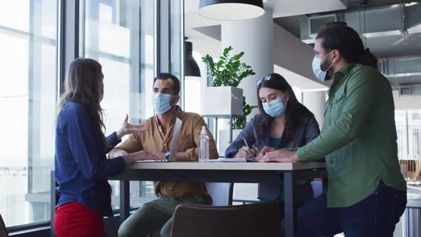 Diverse group of business colleagues wearing face masks sitting at table and talking alt