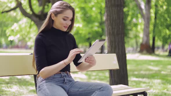 Smiling Beautiful Woman Sitting on Bench in Green Park and Using Tablet alt