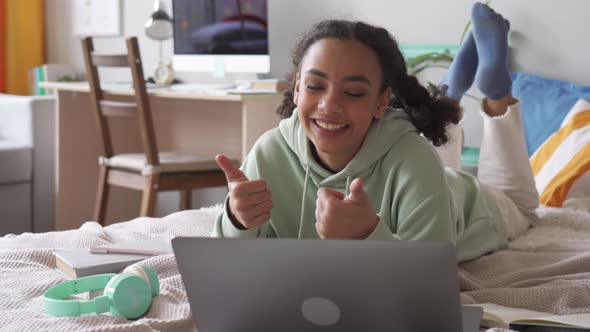 Happy African Teen Girl Talking Video Calling on Laptop Lying on Bed at Home alt