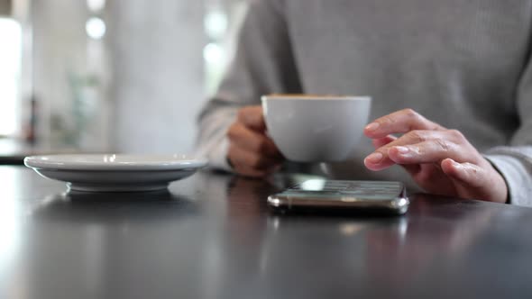 Closeup of a woman touching and scrolling on smart phone screen while drinking coffee