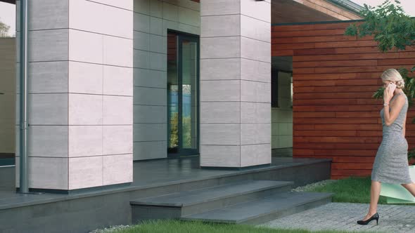 Elegant Woman Talking Mobile Near Luxury House. Beautiful Woman Going with Bags alt