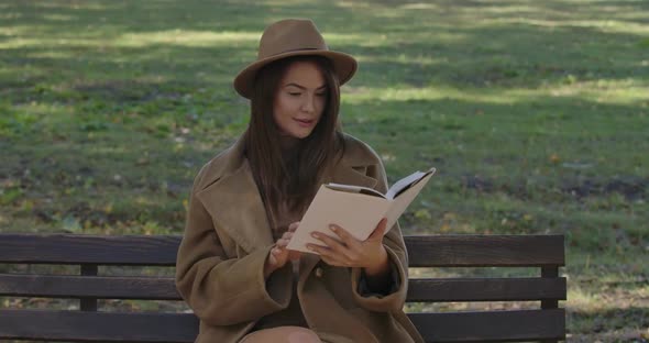 Cute Caucasian Female Student in Elegant Brown Clothes Sitting on the Bench and Turning Book Pages alt