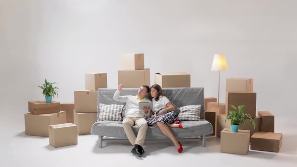 Happy Renters Owners Sit on Sofa Use Digital Tablet on Moving Day in New House Among Cardboard Boxes