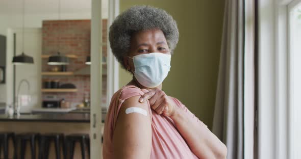 Happy african american senior woman in face mask showing plaster on arm after covid vaccination alt