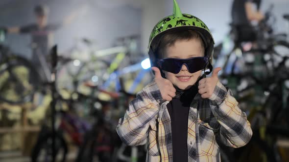 Funny Kid with Sunglasses and Helmet Shows Thumbsup in Shop