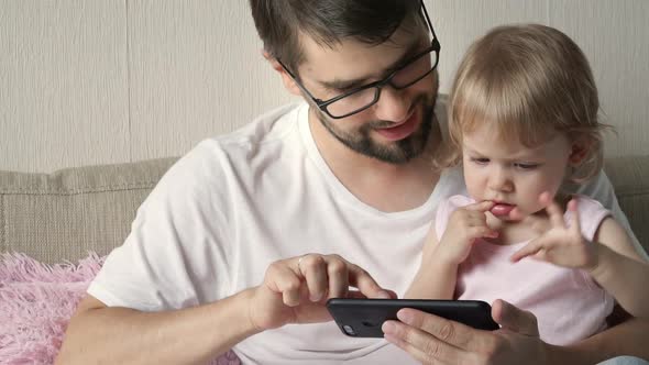 Closeup of Father with Little Daughter Watch Cartoons on Mobile Phone Sitting on Sofa