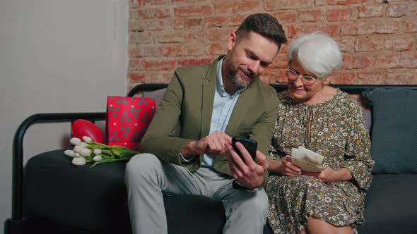 Vivacious Old Greyhaired Woman Gladly Thinks About Past and Shows Her Adult Grandson Old Photographs alt