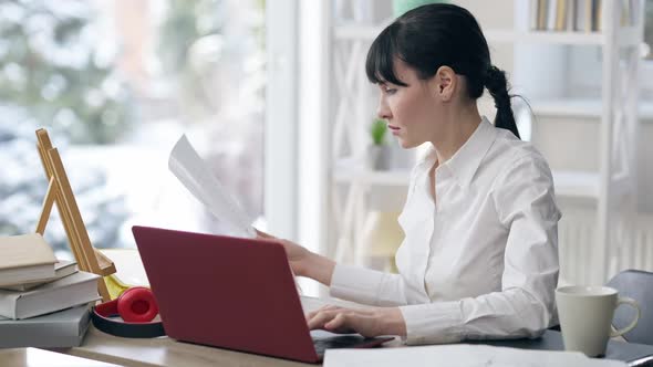 Side View of Confident Focused Young Businesswoman Looking at Documents and Typing on Laptop alt