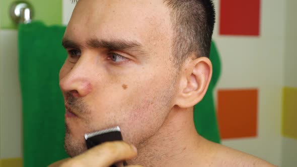 A Caucasian Man Shaves with an Electric Razor Trimmer at Home in the Bathroom alt