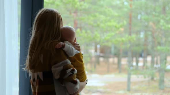 woman her baby in her arms standing at the window with a view of the forest