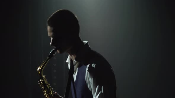 Silhouette a Young Stylish Guy Plays the Golden Shiny Saxophone on Stage. Dark Studio with Smoke and alt