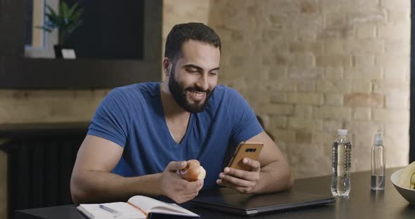 Handsome Young Middle Eastern Man Surfing Internet on Smartphone and Eating Apple. Portrait of Happy alt