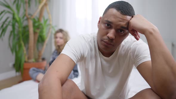 Portrait of Stressed Sad African American Young Man Looking at Camera Sitting on Bed with Blurred alt