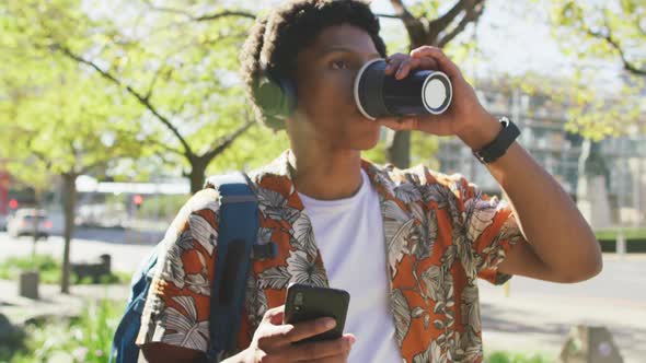 Happy african american man in city, using smartphone and headphones, drinking coffee in street alt