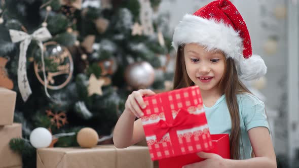 Closeup Portrait of Astonished Girl in Santa Claus Cap Opening Gift Box Having Positive Emotion alt