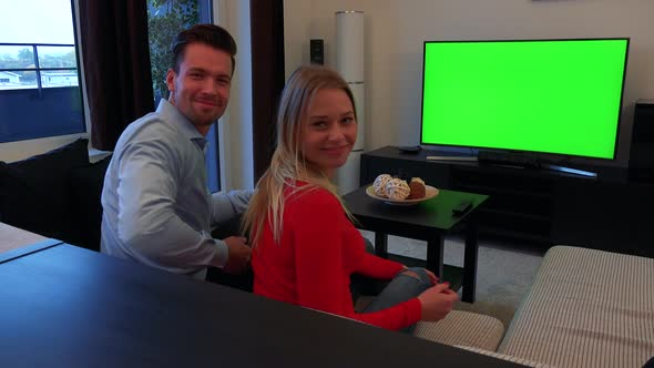 A Couple Watches a TV with a Green Screen in a Living Room, Turns To the Camera and Shows Thumbs Up alt