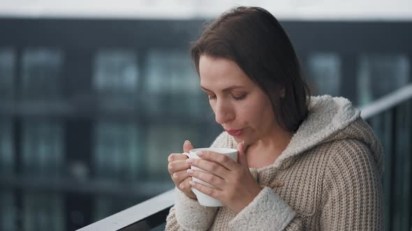 Woman Stays on Balcony During Snowfall with Cup of Hot Coffee or Tea alt