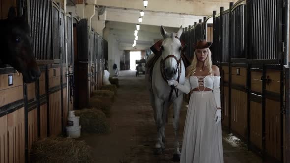 Romantic Woman with Cowboy Hat on Head is Leading White Horse in Stable Holding Reins alt
