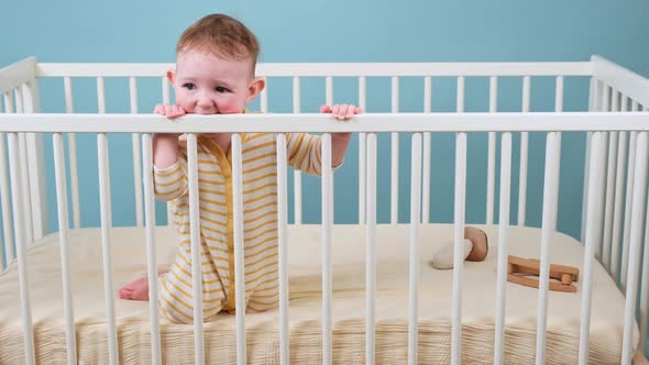 Sad toddler baby boy in the crib gnawing a wooden bars, blue studio background