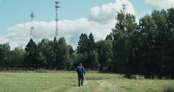 Hiker with Backpack Walks Across Field Against the Background of 5G Radio Towers