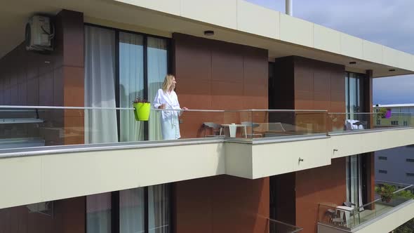 Drone view of woman in white coat with cup of coffee, on hotel balcony alt