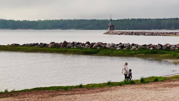 Mother with Child Walks on the Beach Against the Background of Old Chapel Stands on a Stone alt