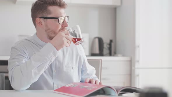 A Young Stylish Man with Glasses in the Morning at Breakfast Reads a Magazine and Drinks Tea