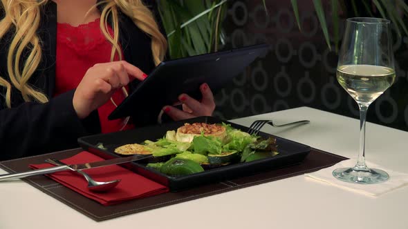 A Woman Sits at a Table in a Restaurant and Works on a Tablet, a Salad in Front of Her alt