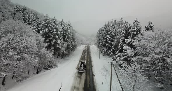 Aerial view of snowy forest and highway while snow cleaner car removes snow from the road alt