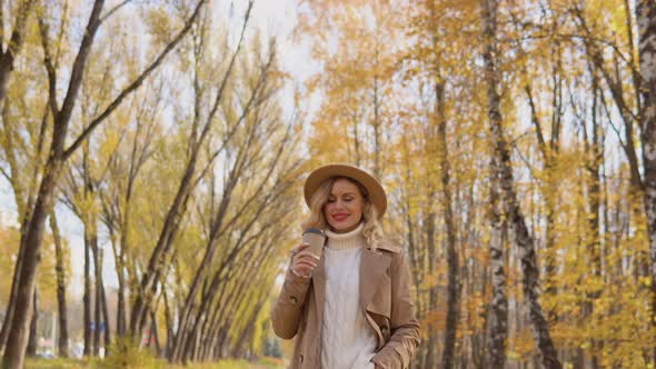 Young Woman in a Brown Coat and Hat Walks in the Autumn Park and Drinks Coffee alt