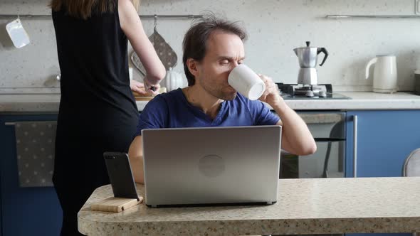 Man Freelancer Starts to Work on Laptop While His Wife Preparing Breakfast in the Kitchen