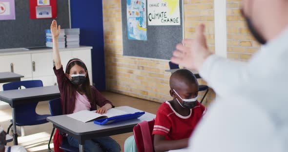Diverse schoolchildren sitting in classroom raising hands during lesson, all wearing face masks alt