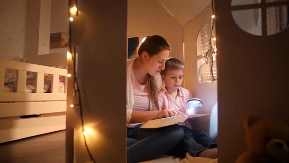 Panning Shot of Young Smiling Woman Reading Story Book To Her Little Son at Night While Sitting on alt