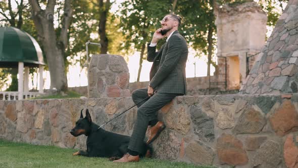 Wealthy Businessman in a Business Suit Is Talking on the Phone and Smiling with a Doberman Dog alt