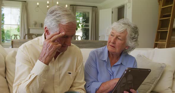 Stressed caucasian senior couple smiling while using digital tablet sitting on the couch at home alt